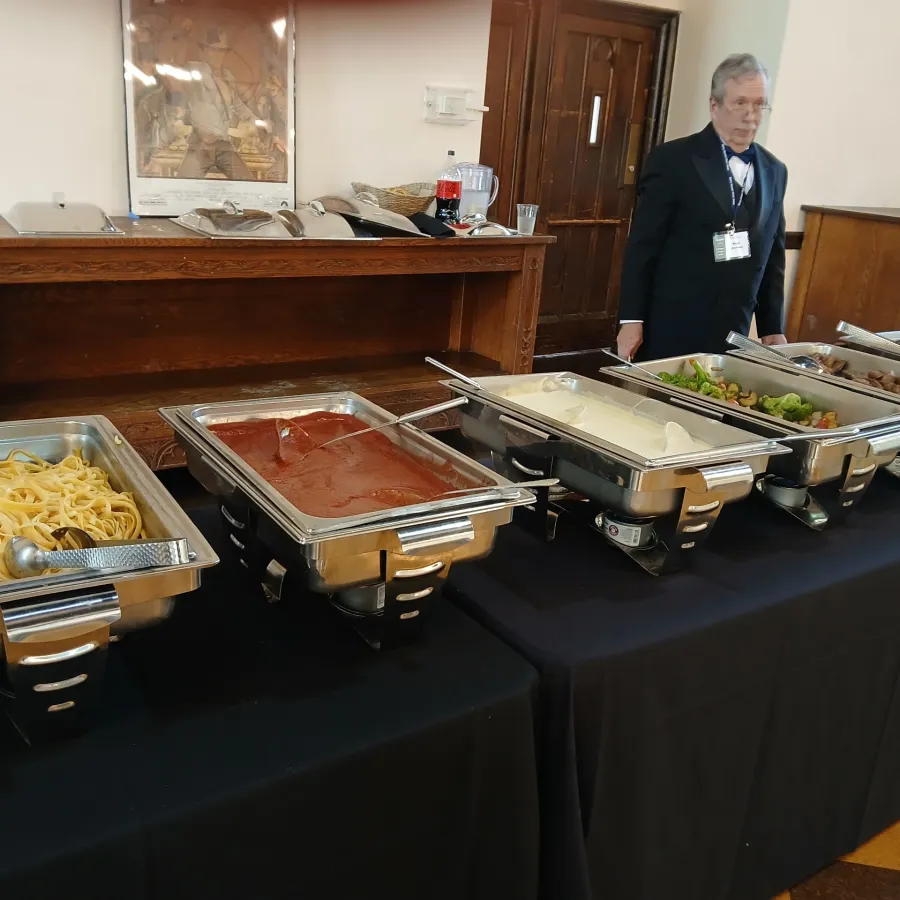 Buffet table with chafing dishes containing pasta, sauces, and vegetables, attended by a suited server.