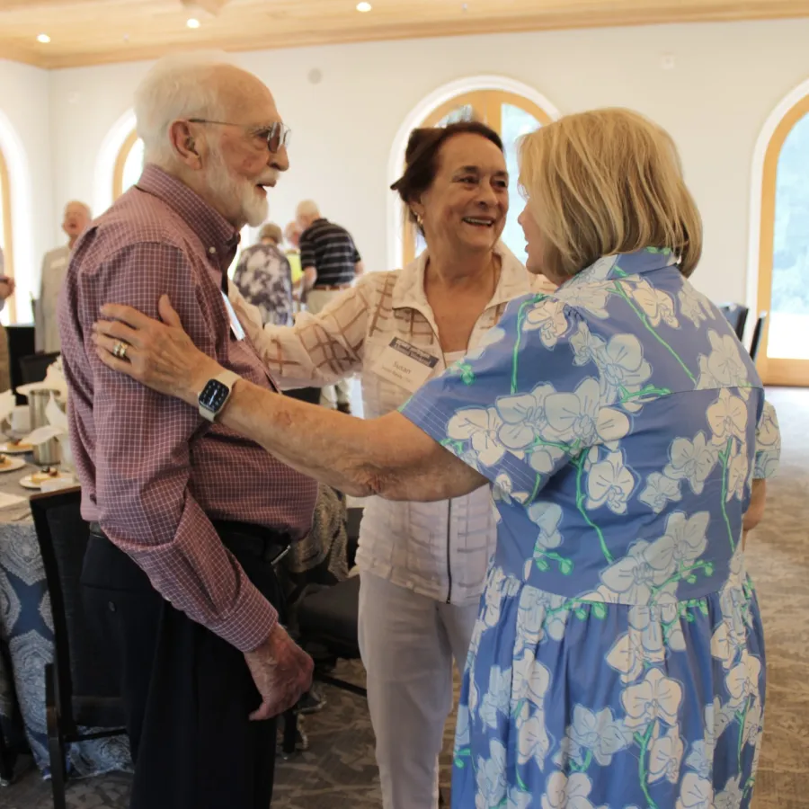 Three seniors happily dancing together in a bright room with wooden ceiling and carpeted floor.