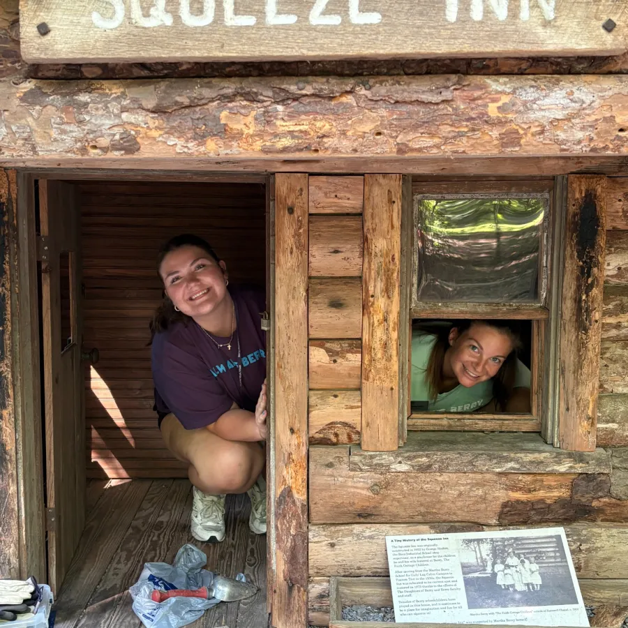 Two people smiling inside a rustic wooden cabin with a sign reading Squeeze Inn outside.