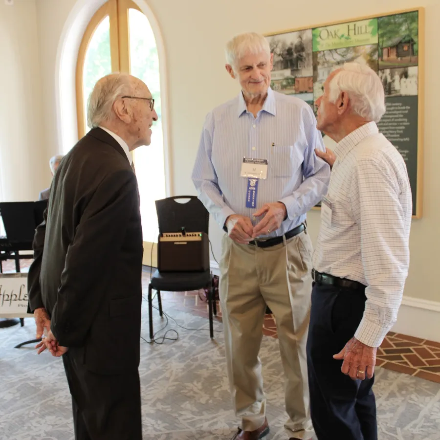 Three elderly men engaged in conversation in a well-lit room with carpet and wooden window frames.