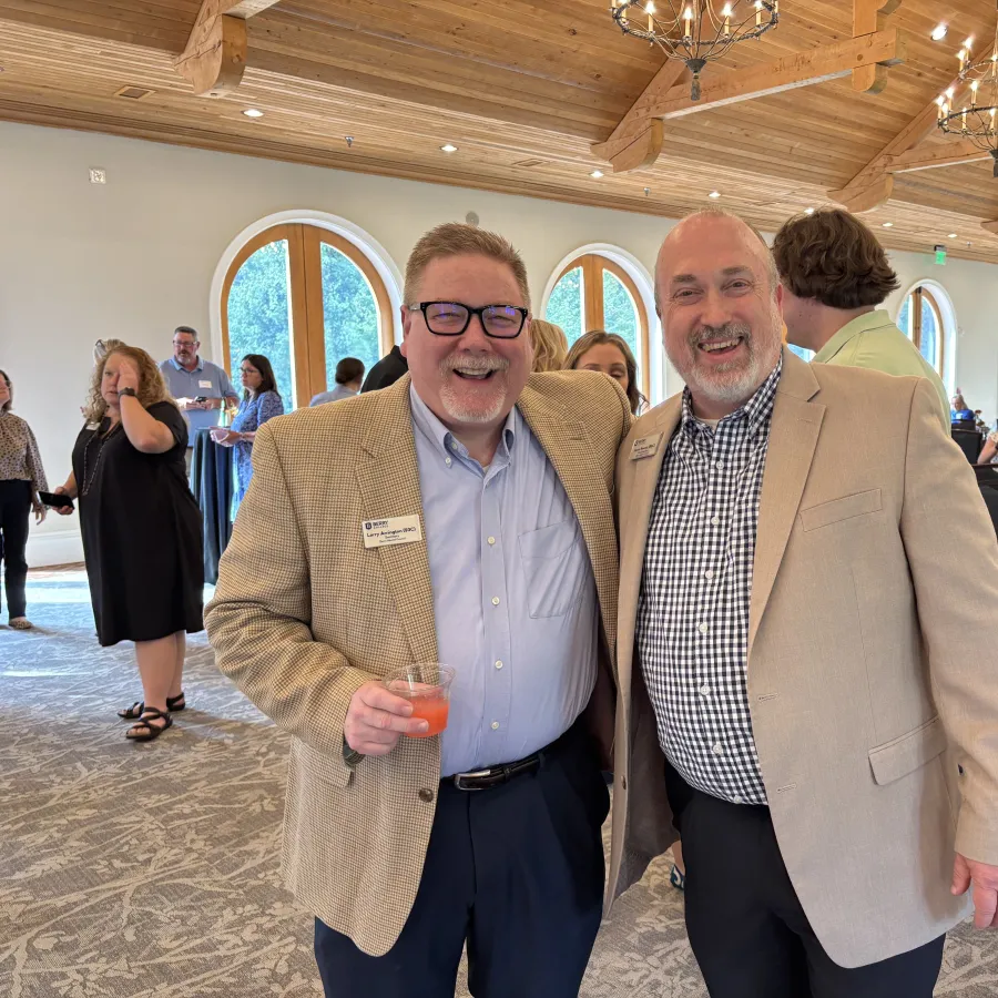 Two men in blazers smiling and posing at a social event inside a wood-ceilinged room with chandeliers and arched windows.