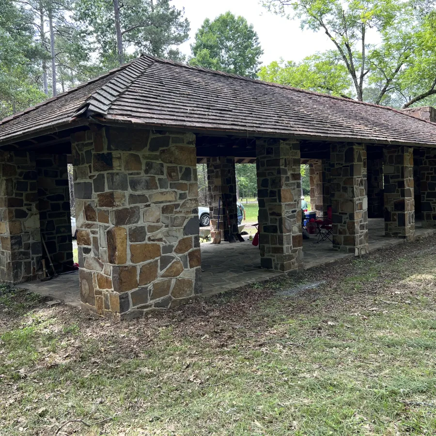 Stone pavilion with wooden shingle roof supported by stone pillars in a forested park area