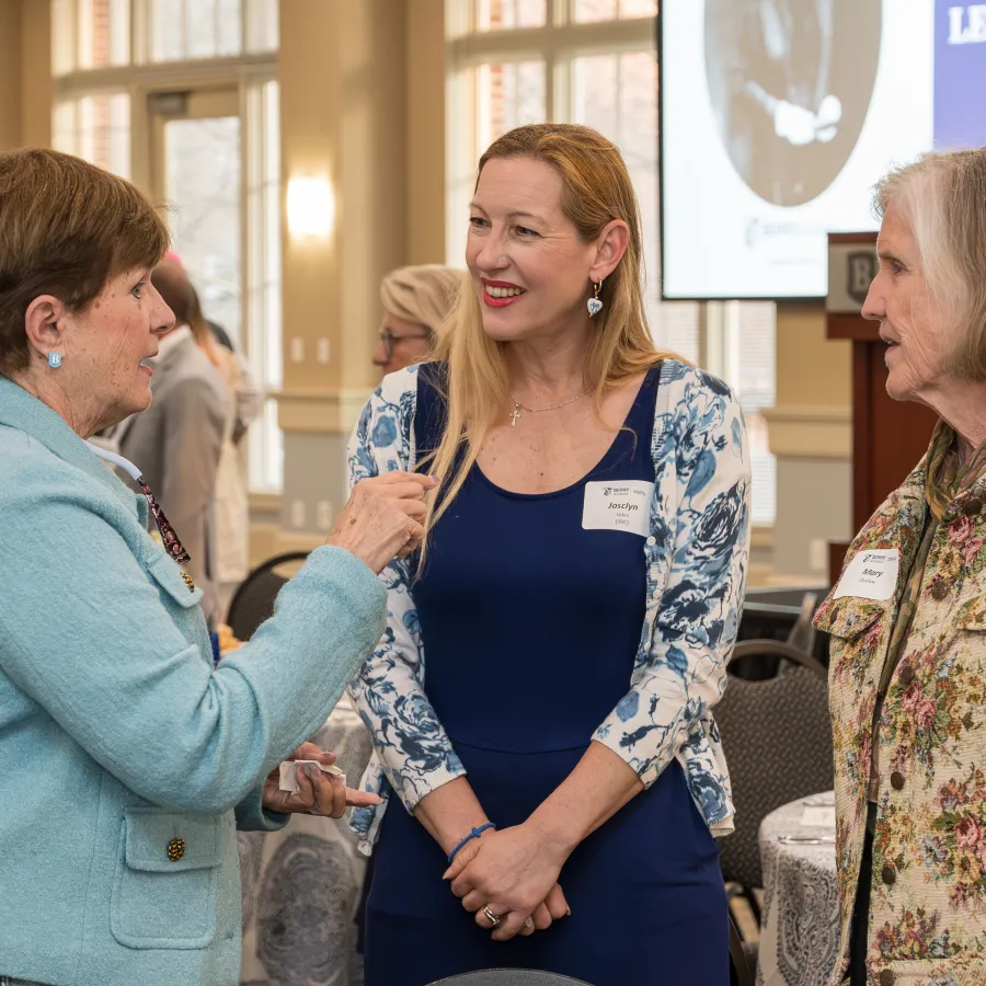 Three women engaged in conversation at a formal Legacy Dinner event in a bright banquet room
