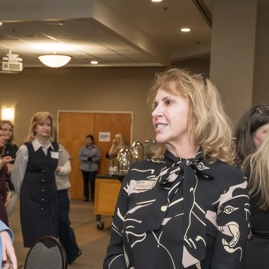 Two professional women engaged in conversation at a networking event with other attendees in the background.