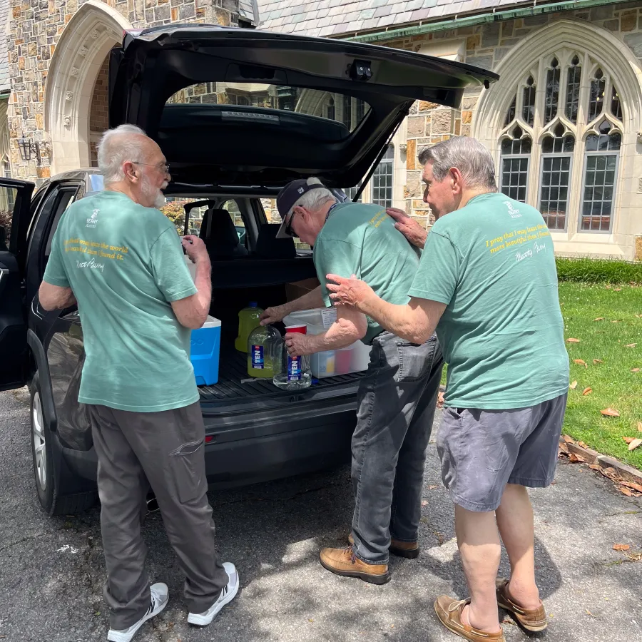 Three elderly men in matching green shirts unloading supplies from a car trunk near a stone building with arched windows.