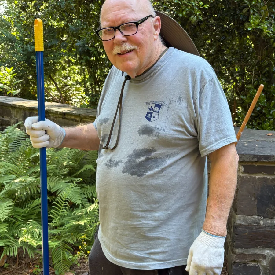 Elderly man wearing gloves and holding a rake in a garden with greenery and stone path background.
