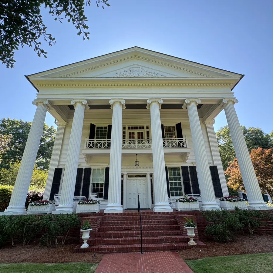Historic white mansion with large columns and brick walkway surrounded by greenery under clear sky
