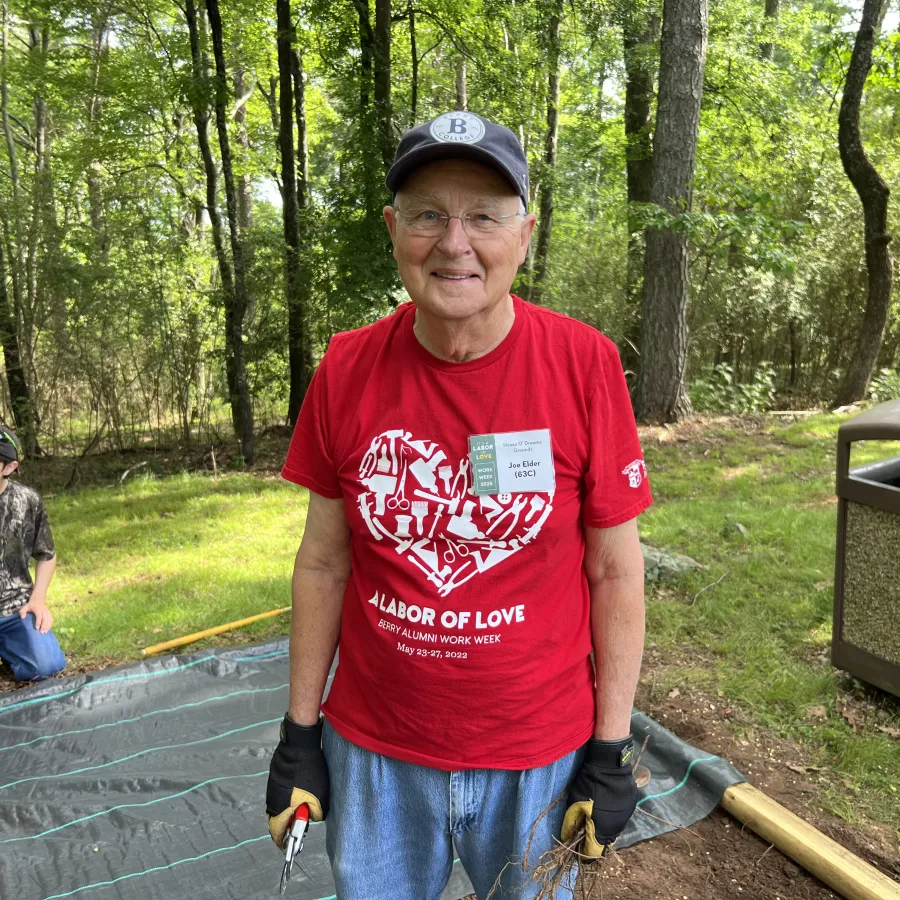 Senior man wearing a red Labor of Love shirt and gloves, standing outdoors with trees and a young boy in the background.