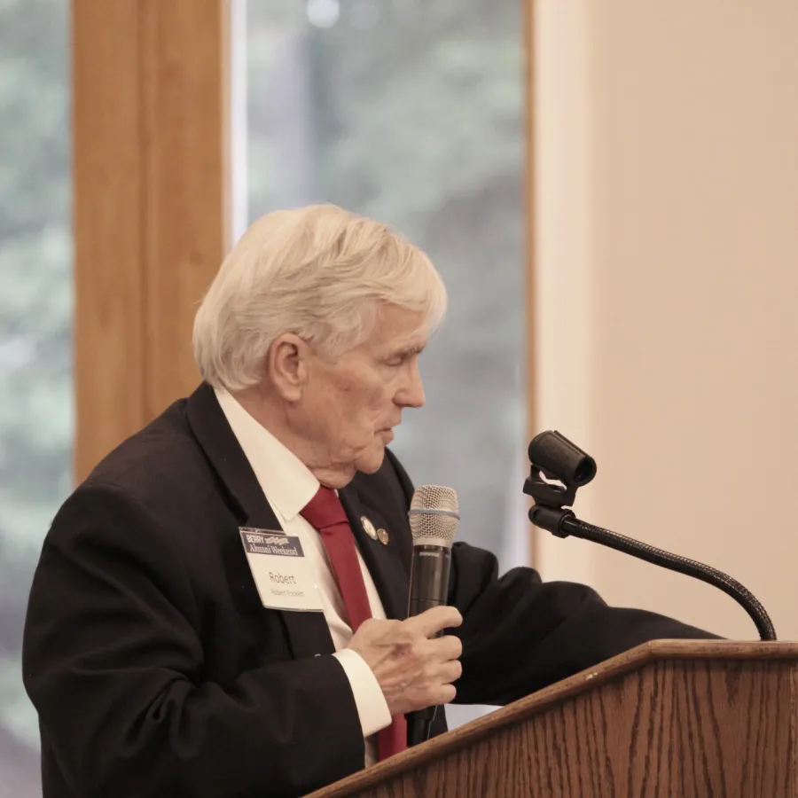 Older man in black suit and red tie speaking at podium with microphone in formal setting