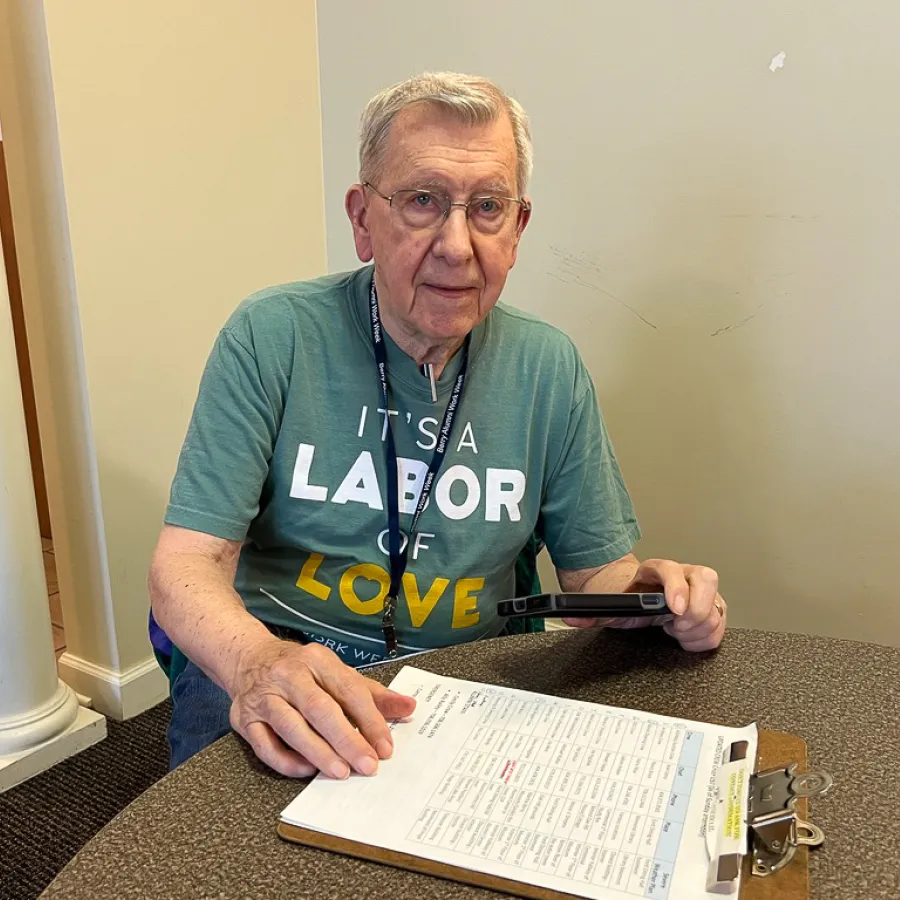 Elderly man wearing a green t-shirt reading 'It's a Labor of Love' using a phone and pointing at a clipboard on a table.
