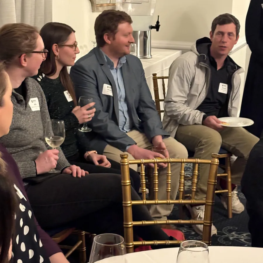 People seated and socializing at a formal event with drinks and food on a white tablecloth-covered table.