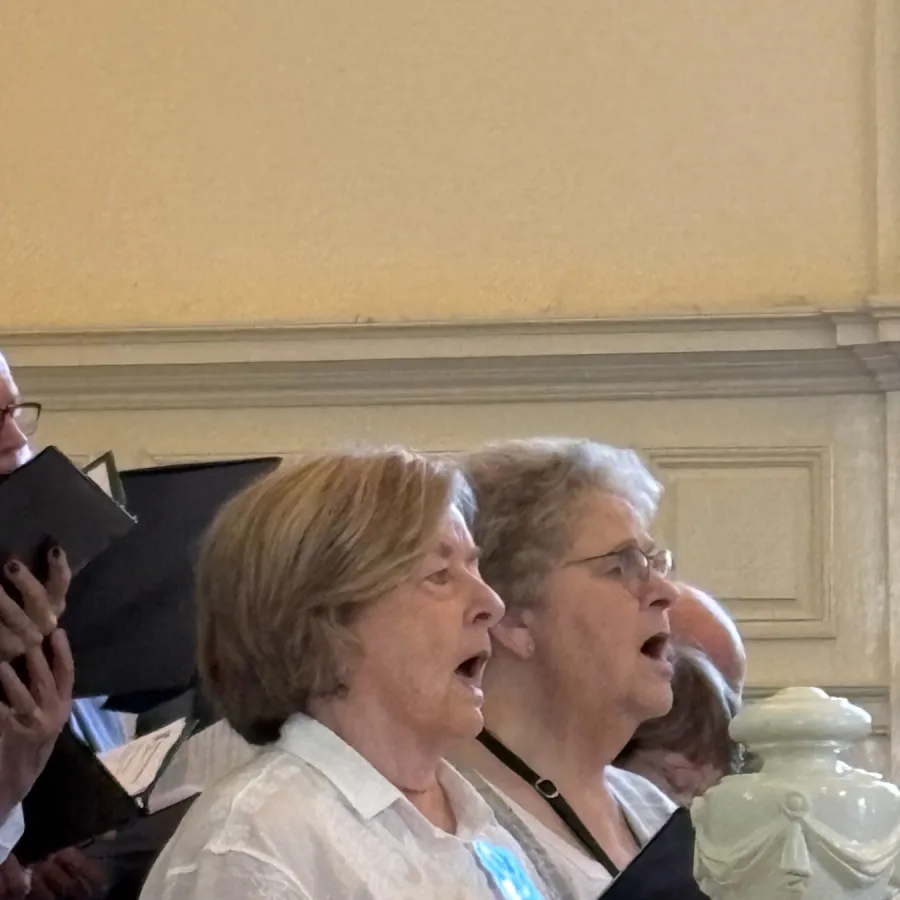 Group of elderly women singing together indoors near ornate white railing in a formal setting