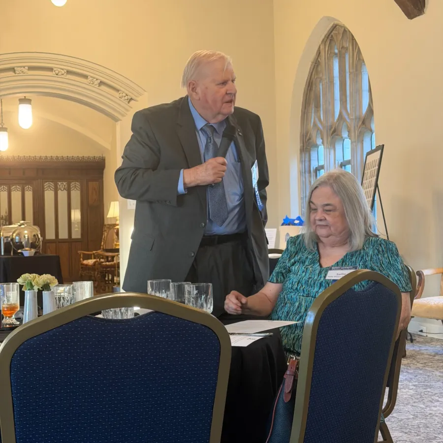 Elderly man in suit conversing with elderly woman seated in a church or formal setting with arched windows.