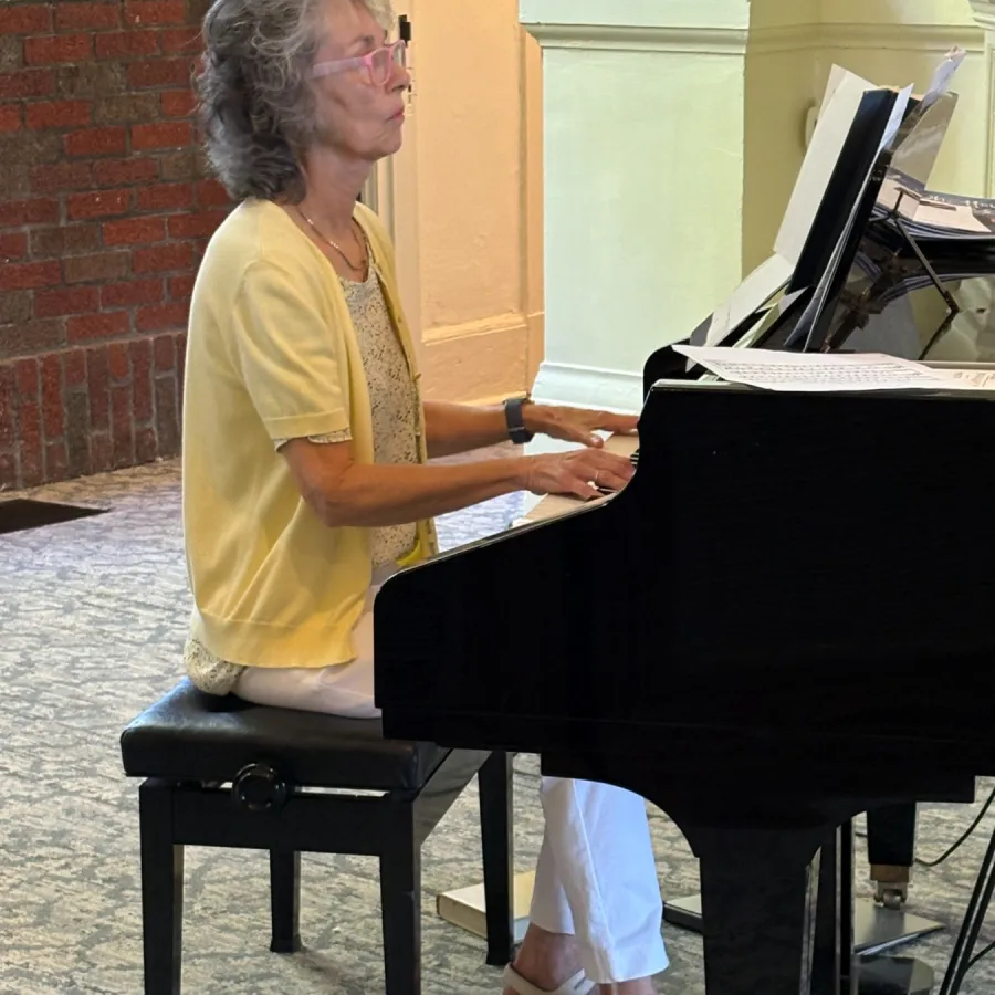 Elderly woman wearing a yellow blouse playing a black grand piano seated on a piano bench indoors.