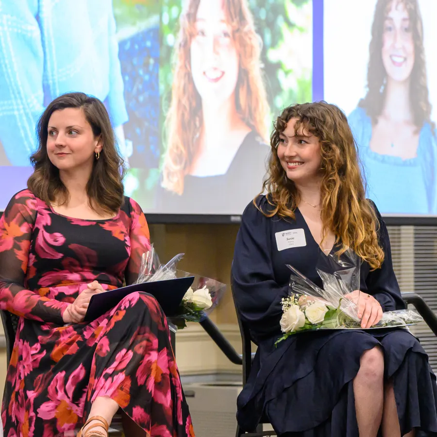 Four women seated indoors at an event holding flowers, one speaking into a microphone with a projected image behind them