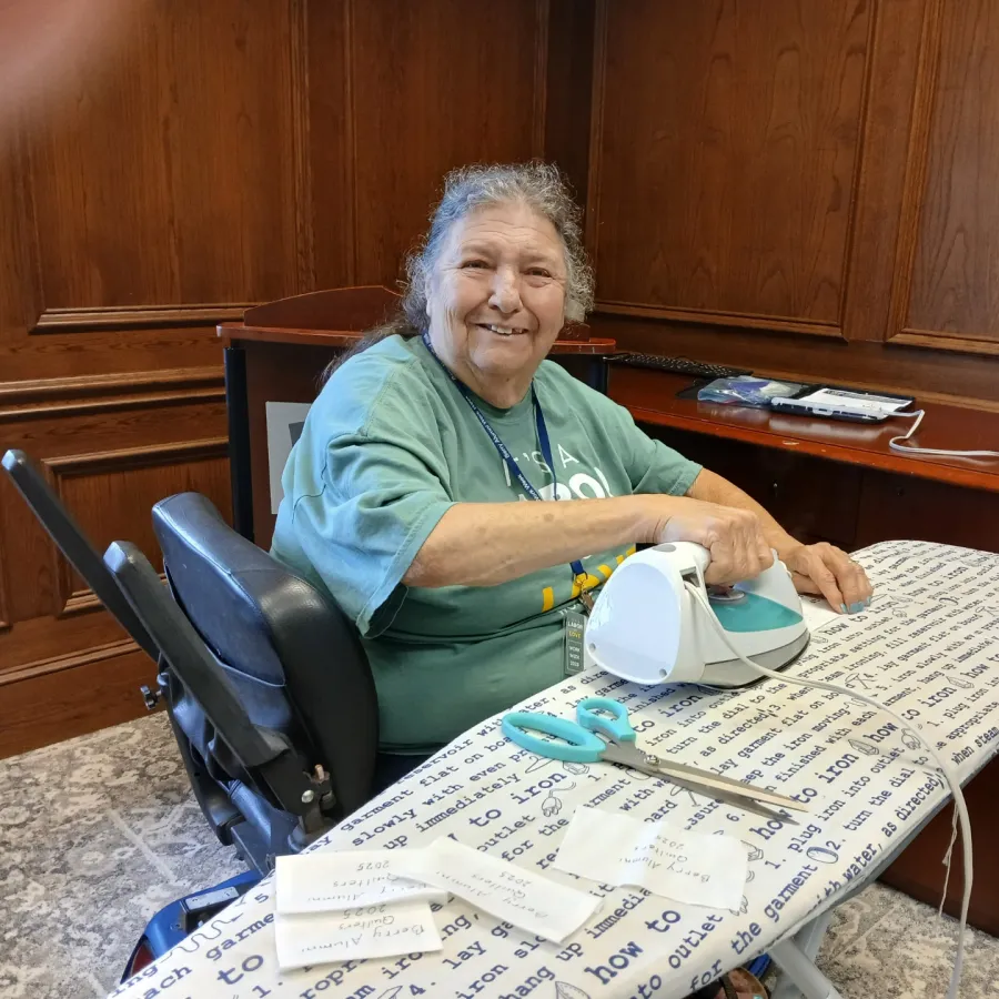 Elderly woman smiling while using a small iron on fabric pieces at an ironing board in a wood-paneled room.