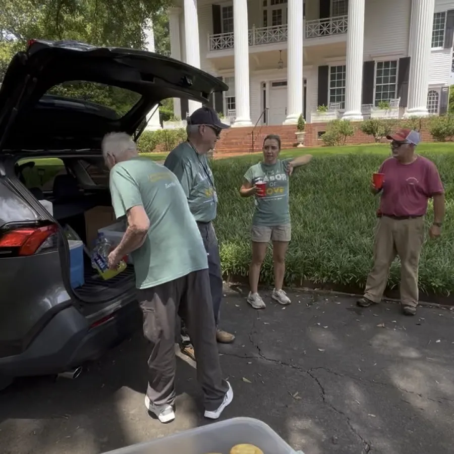 Group of adults chatting near a car with snacks in front and a large white columned building in the background