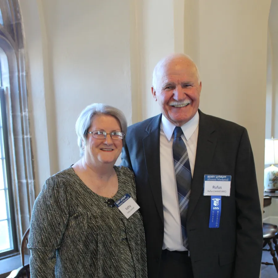 Smiling older man and woman wearing name tags posing indoors near window with blinds and tile floor.