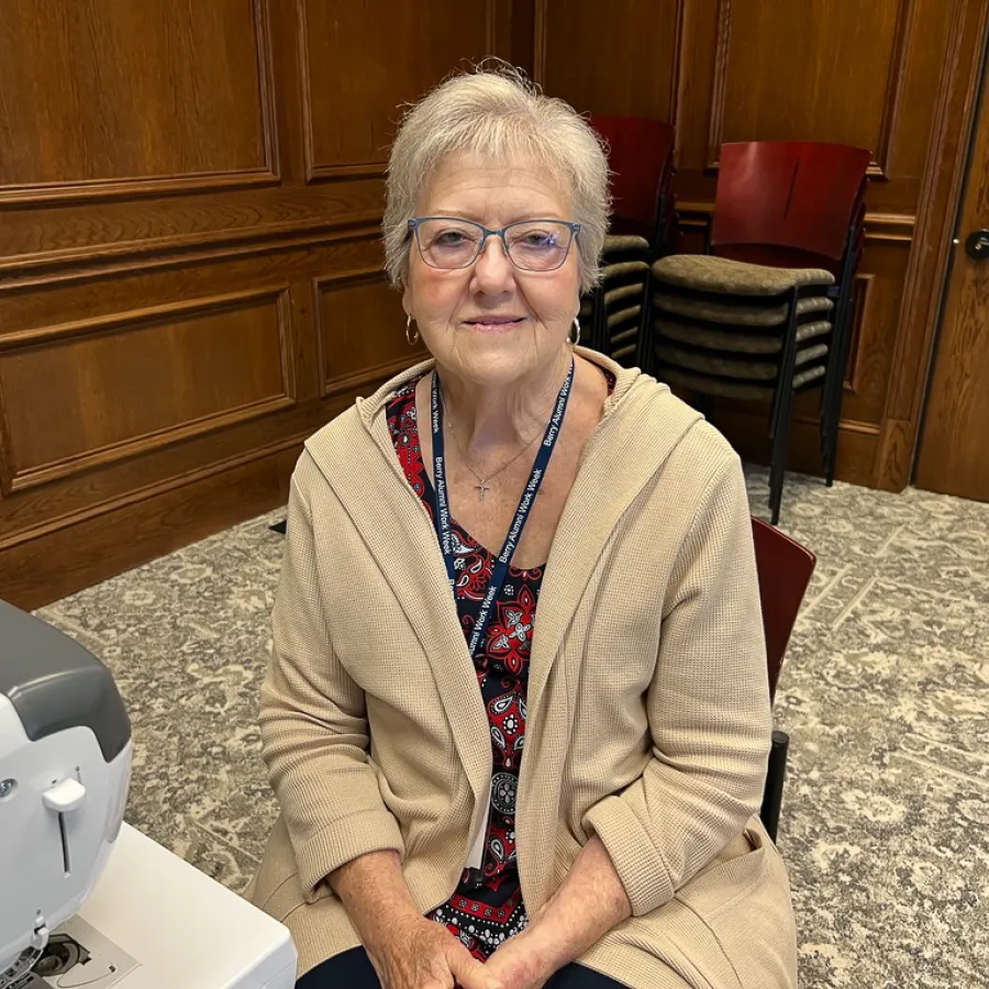 Elderly woman with glasses and beige cardigan sitting at sewing machine in a wood-paneled room.