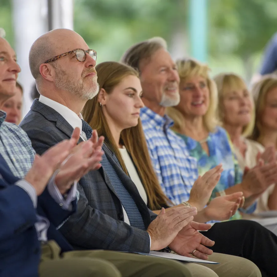 Diverse adults seated outdoors clapping and attentively watching an event on a sunny day.