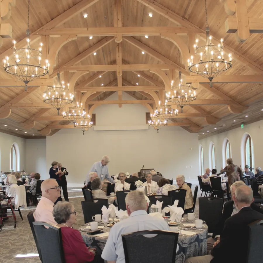 Seniors dining in a spacious room with wooden vaulted ceiling and chandeliers during a social event.