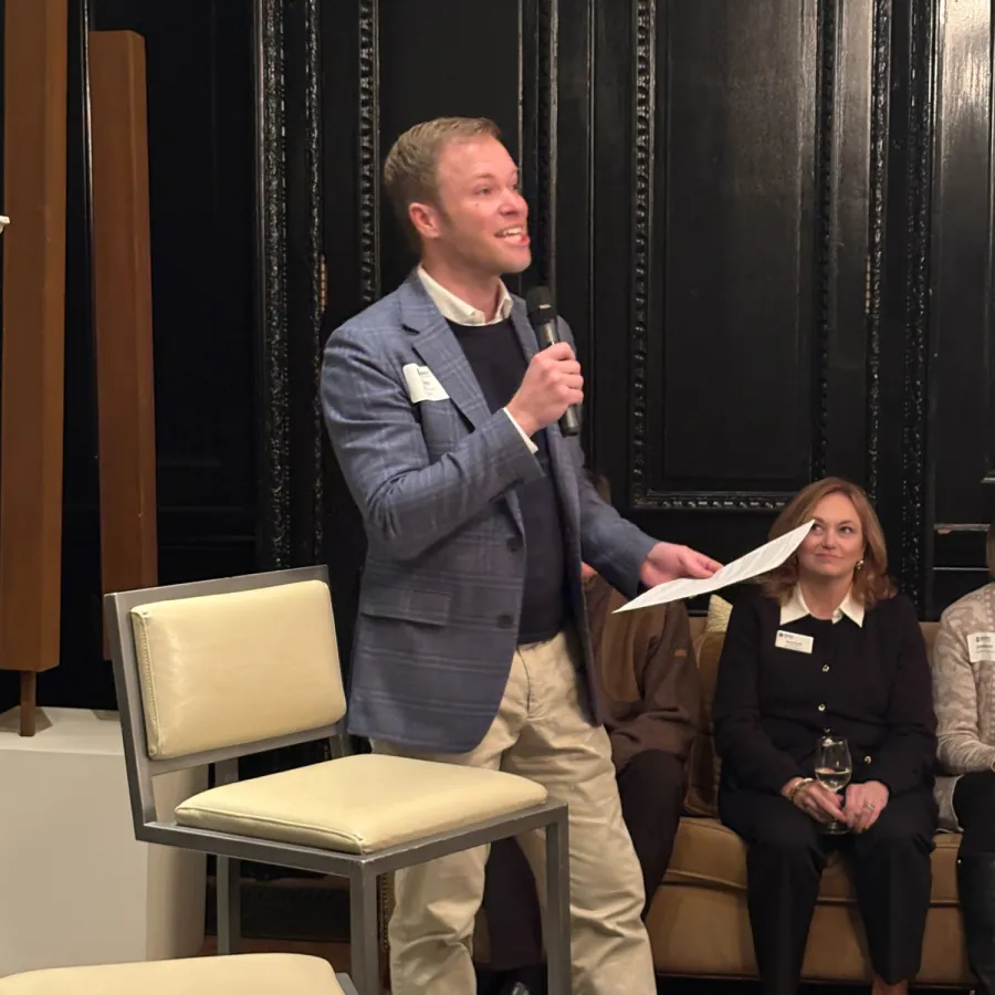 Man in blazer speaking into microphone at event while seated audience listens attentively indoors