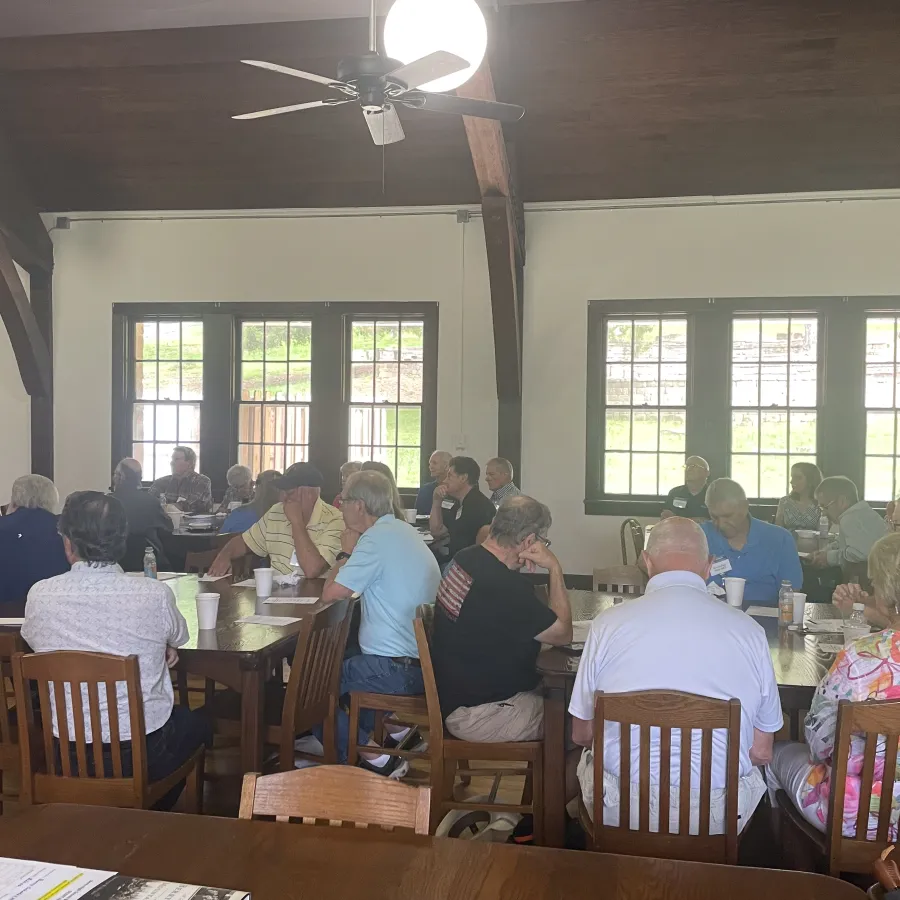 Group of elderly people seated at tables in a bright room with wooden ceiling and large windows during a meeting.