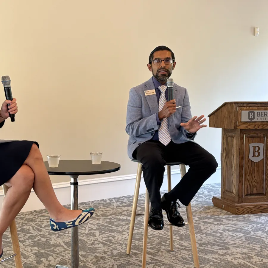 Two professionals holding microphones engaging in a discussion on stools with a small table and podium in conference room.