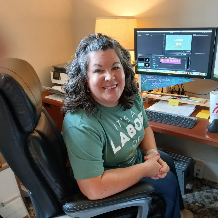 Smiling woman in green labor day shirt sitting in office chair at desk with dual monitors and Chick-fil-A cup