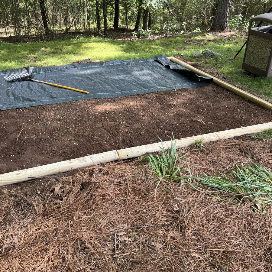 Garden bed area prepared with wooden frame, soil, rake, tarp, and nearby tools in a backyard setting.