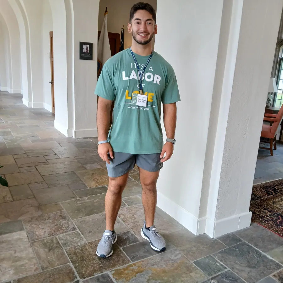 Young man standing in a bright hallway wearing a green labor of love t-shirt and shorts with a conference badge.