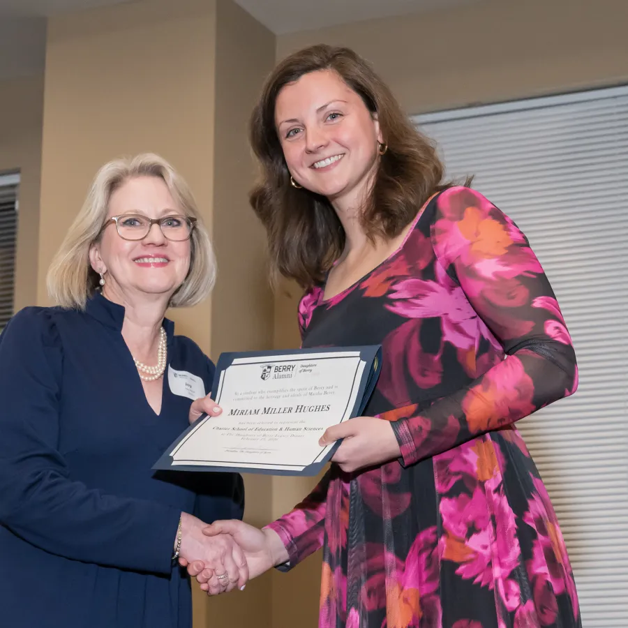 Two women smiling while shaking hands as one receives a certificate award indoors at an event.