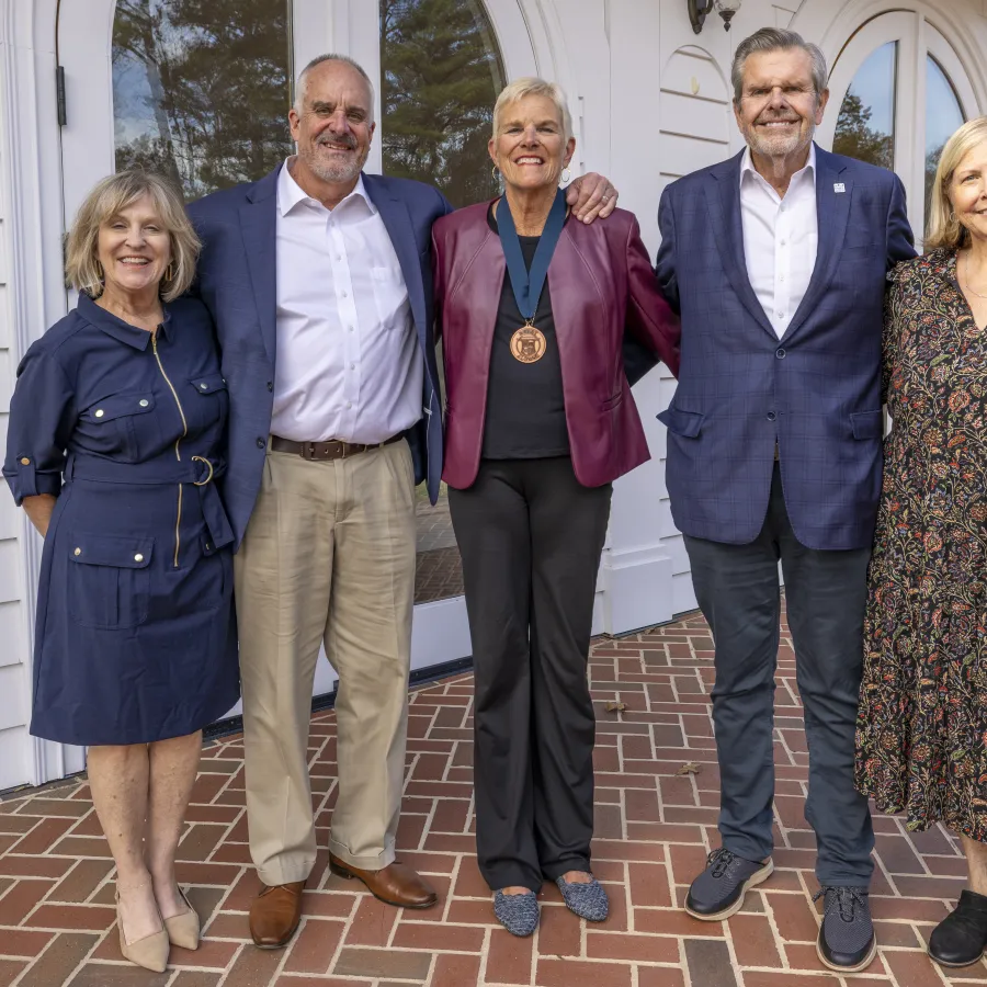 Five adults standing on a brick patio in front of white building with arched windows, one wearing a medal.