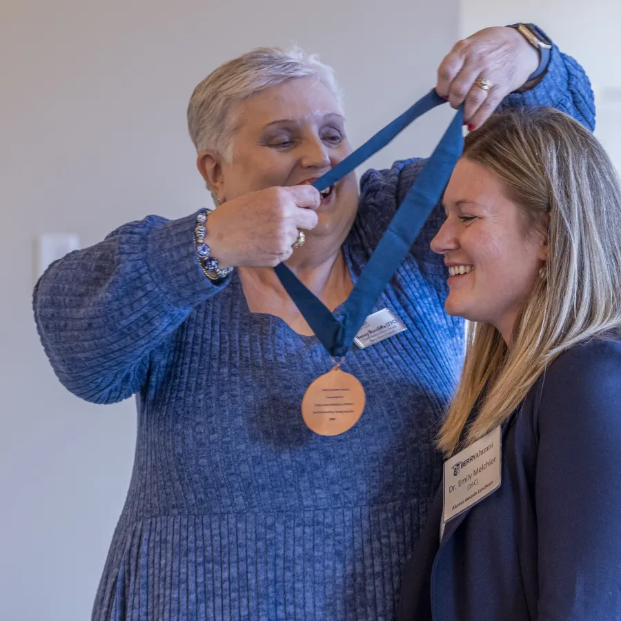 Older woman awarding a medal to a smiling younger woman at an indoor event with name badges.