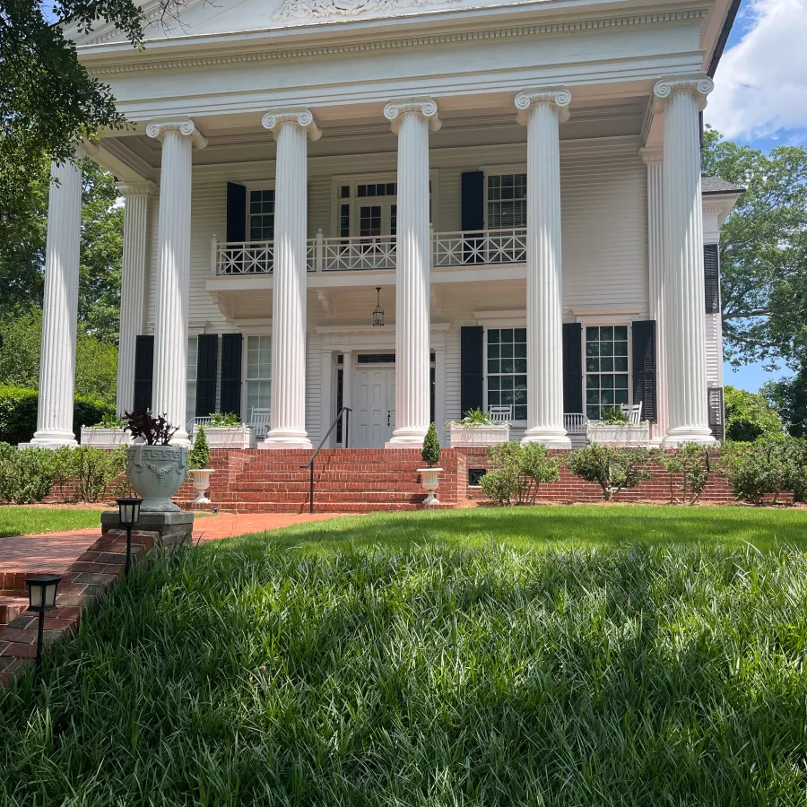 Elegant white Southern mansion with tall columns, black shutters, brick steps, and lush green lawn under blue sky.