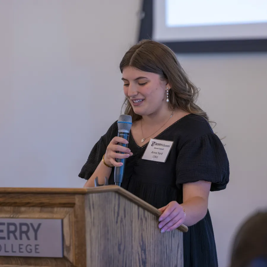 Young woman speaking at Berry College podium holding microphone, delivering a presentation in a formal setting.