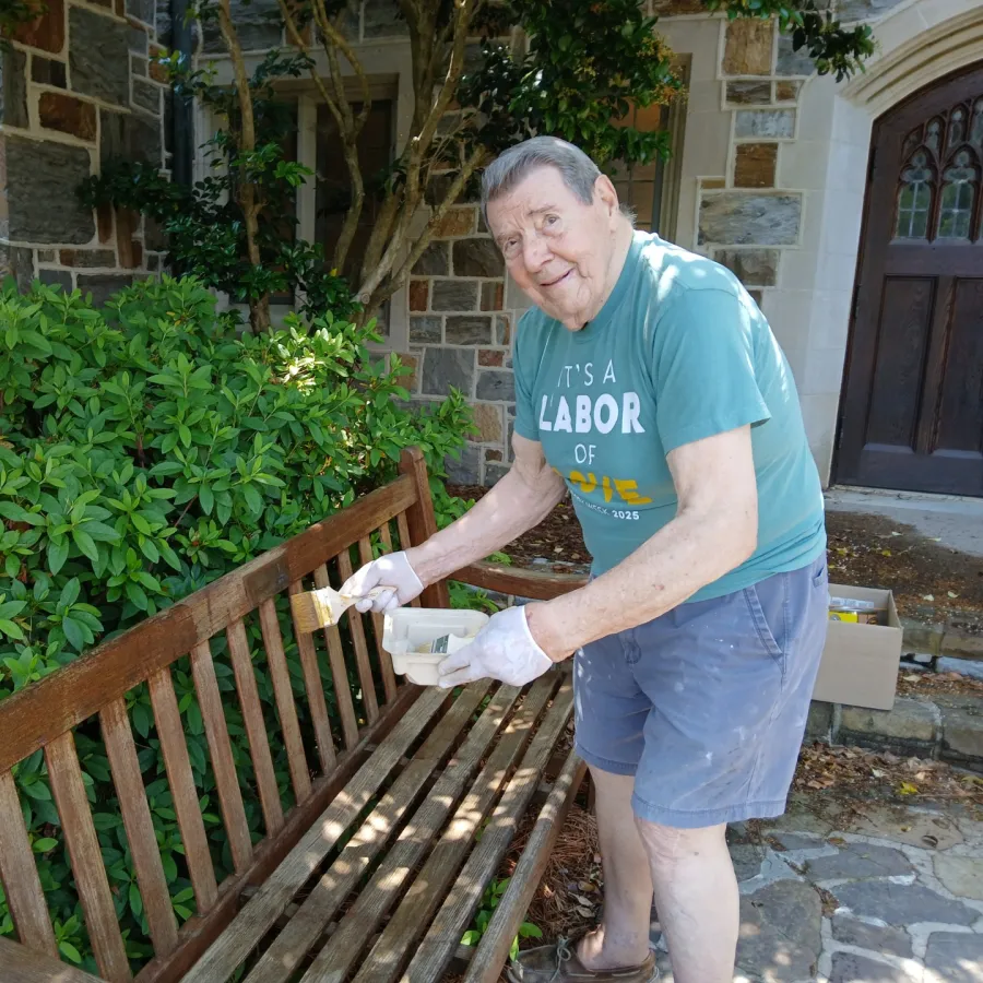 Elderly man wearing gloves painting a wooden bench outdoors near stone building and greenery.