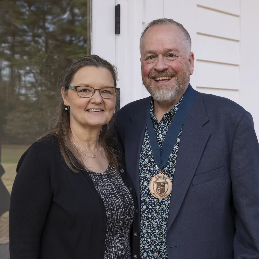 Smiling man wearing a medal and woman standing together outside near a glass door on a sunny day