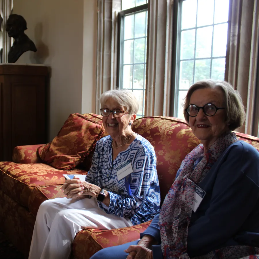 Three elderly women sitting and smiling in a warmly lit living room with vintage furniture and large windows.