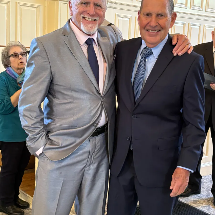 Two older men in suits smiling and posing together indoors with a woman seated in the background.