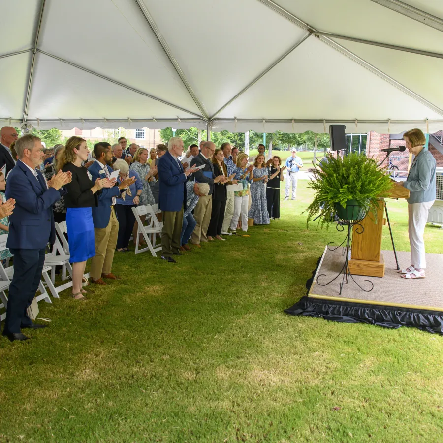 Audience standing and applauding a speaker on stage under a large outdoor event tent on grass.
