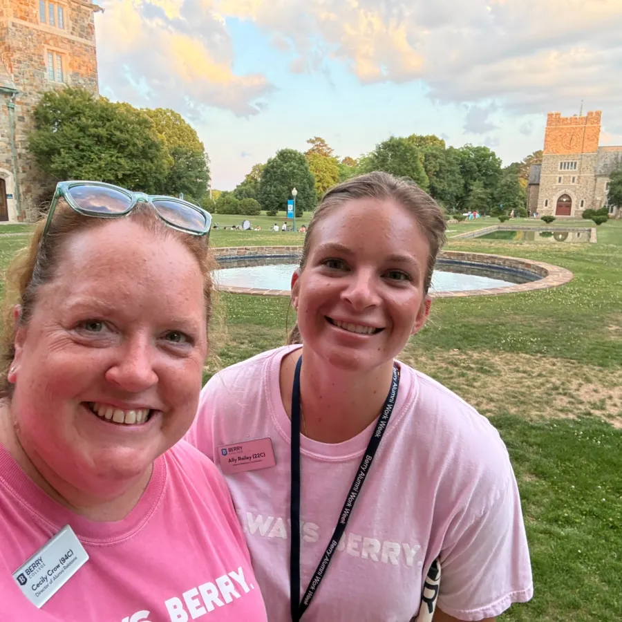 Two women in pink shirts smiling outdoors in front of historic stone buildings and a green lawn with a fountain under a cloudy sky