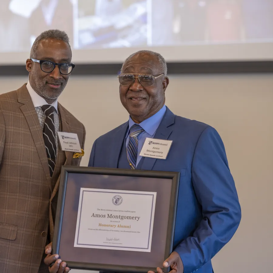 Two men in suits hold a framed honorary alumni certificate at an indoor event with a presentation screen behind them