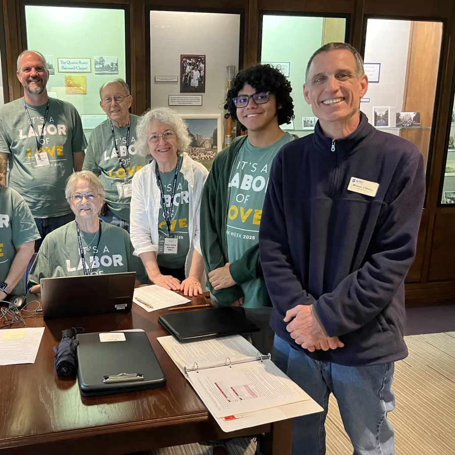 Group of smiling volunteers wearing matching shirts with Labor of Love text gathered around a table with laptops and papers.