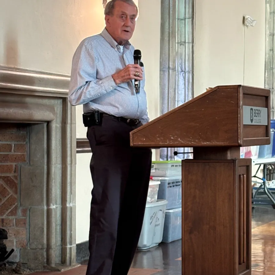 Man in formal attire speaking into microphone at wooden podium in indoor setting with tiled floor.