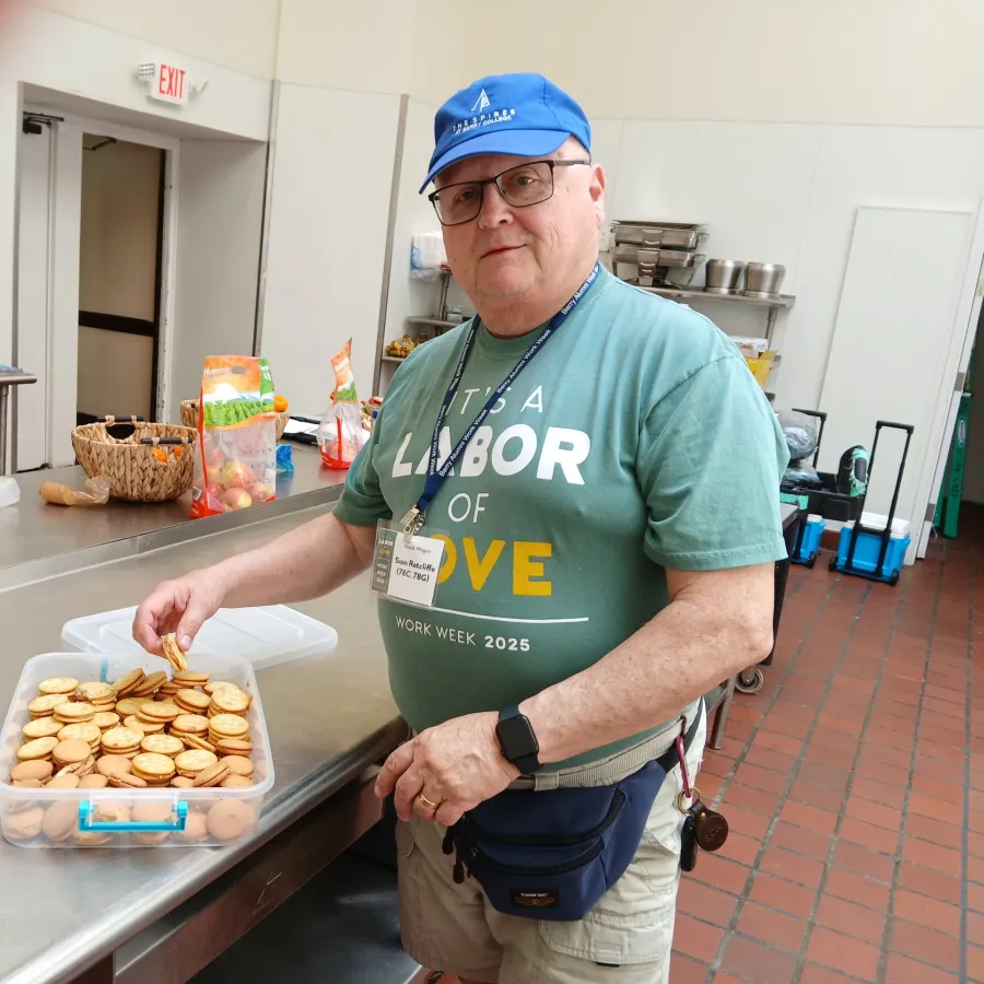 Man in a blue cap and teal shirt holding crackers near a container of snacks in a kitchen with metal counters.
