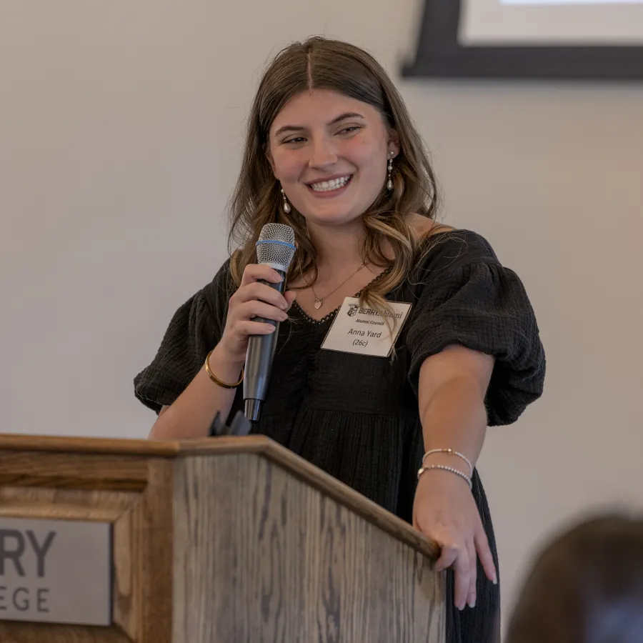Young woman speaking at Berry College podium with microphone, smiling during presentation or speech