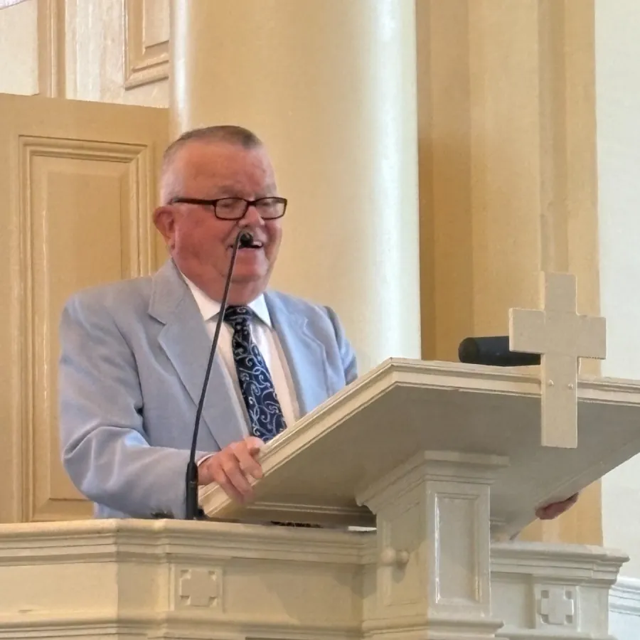 Man in light suit speaking at ornate white church pulpit with a cross and microphone