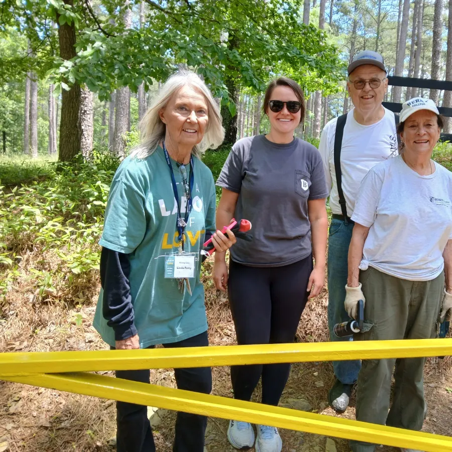 Four volunteers in casual clothing stand outdoors holding tools by yellow caution tape in a wooded area on a sunny day