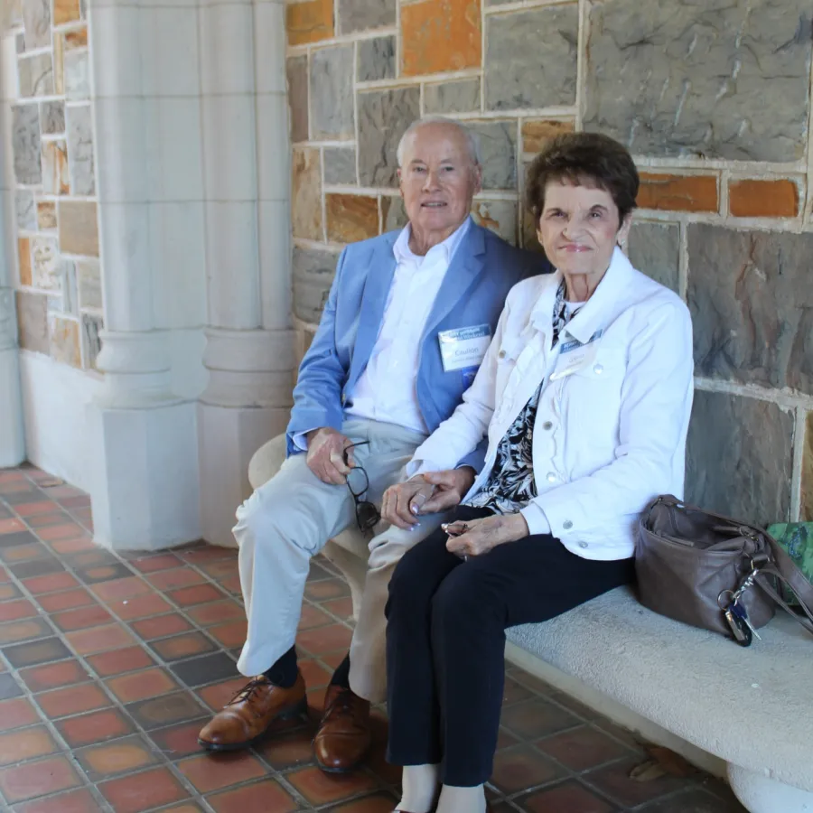 Elderly couple sitting on a stone bench inside a building with tile floor and stone wall background.